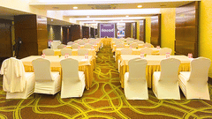 A conference room at Pride Plaza Ahmedabad, featuring rows of tables with white tablecloths, chairs, and a projector screen, suitable for meetings and presentations.